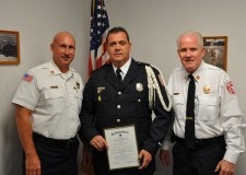 Three men in uniforms; one holds a certificate. An American flag is in the background.