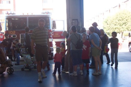 A group of people, including children, are gathered in front of a fire truck inside a garage or fire station.