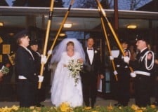 A bride and groom walk under an arch of poles held by uniformed individuals, possibly at a ceremonial wedding exit.