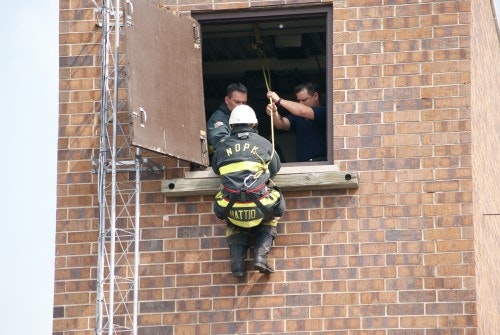 Firefighter rappelling down a brick building, assisted by two people from a window.