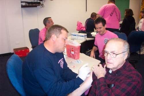 A person receives a vaccine in a clinic setting, with healthcare workers in pink shirts assisting others in the background.