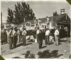 A group of people standing and building a structure outside, near a house with construction materials around.