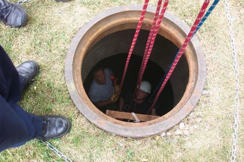 Two people in a manhole, one wearing a helmet, with ropes above, while others stand around the opening.