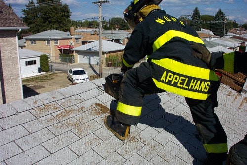 A firefighter in gear is using a tool on a rooftop in a residential area.