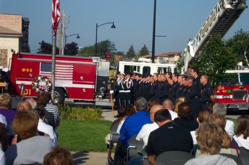 A memorial ceremony with firefighters standing in formation, an American flag on a fire truck, and an audience seated outdoors.