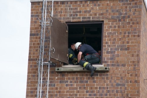 Two firefighters in gear are at a window ledge on a brick building, engaged in an activity or rescue drill.