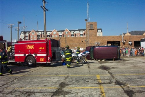 Firefighters and a fire truck at the scene of a vehicle on its side, with bystanders in the background.