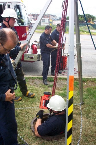 Emergency personnel conducting a rescue drill with equipment around a manhole, one person is descending while others assist.