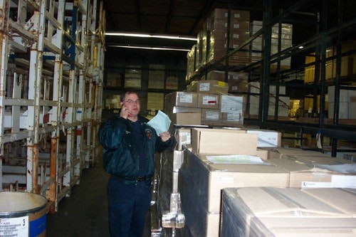 Person in a warehouse using a mobile phone, surrounded by stacked boxes and shelving.