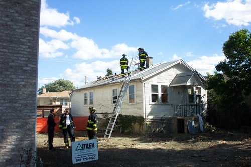 Firefighters on a roof next to a house using a ladder, with a construction company sign in the yard.