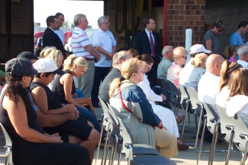A group of people seated and standing, attending an event or ceremony outdoors.