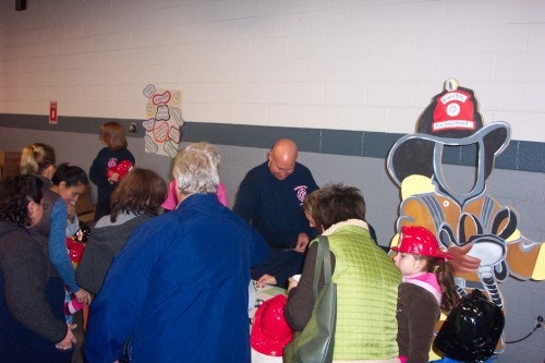 A group of people gather around a table with a firefighter. Children have red helmets, and there's a large firefighter cutout displayed.