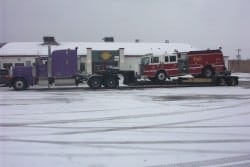 A semi-truck is hauling a red fire truck on a flatbed trailer in a snowy parking lot.