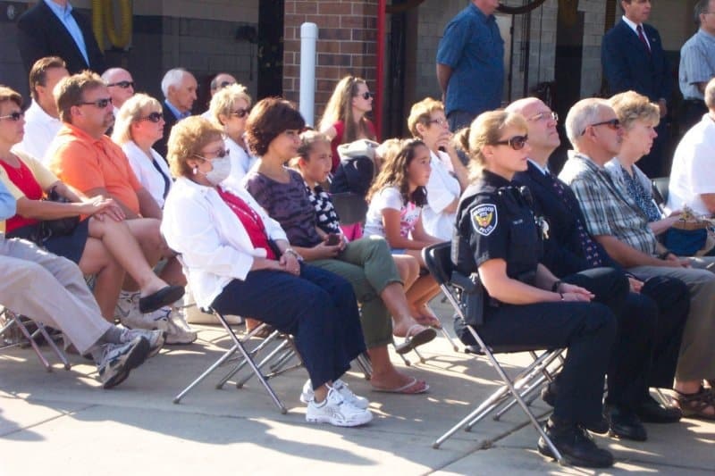 A group of people seated outdoors, including a police officer and a masked woman, participating in an event or ceremony.