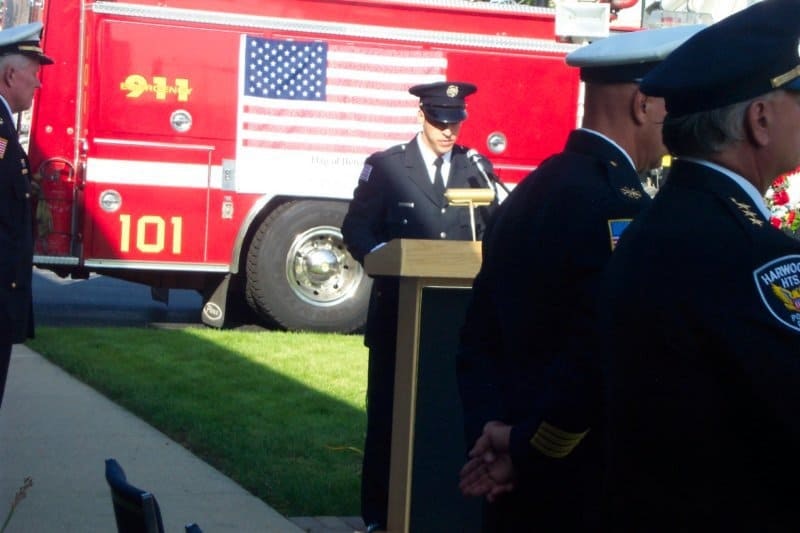 A firefighter speaks at a podium with a fire truck displaying the American flag in the background and uniformed officers present.