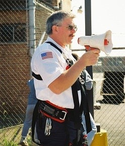 A person wearing a harness and holding a megaphone by a fence, with a U.S. flag patch on their shirt.