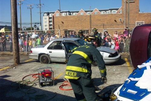 Firefighters demonstrate vehicle extraction in front of a crowd, with a car and emergency equipment visible.