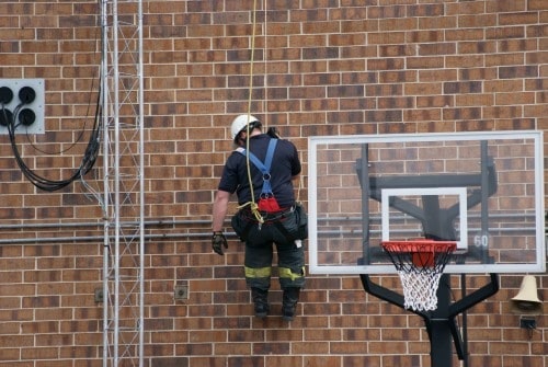 A person in safety gear is secured with ropes, working on a brick wall near a basketball hoop.