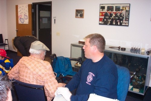 A man in a fire department shirt talks to an older man in a cap. There's a display case and a firefighting photo on the wall.