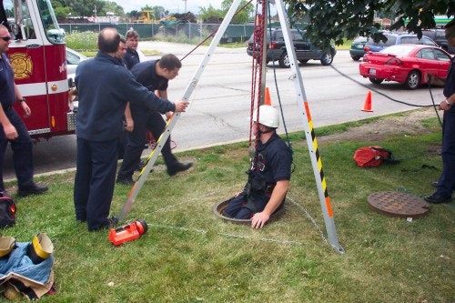 Firefighters conduct a rescue drill with a tripod setup around a manhole; one person descends into the opening.
