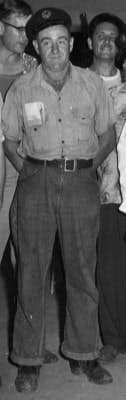 A man in a uniform stands with hands in pockets; others partially visible behind him.