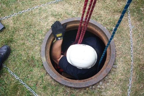 A person with a helmet is entering or exiting a hole in the ground, using ropes for support, surrounded by grass and chains.