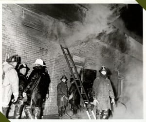 Firefighters in protective gear, smoke rising from a building, ladder propped against the wall, nighttime scene.
