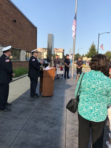 A group of people, including uniformed individuals, gathers outdoors near a podium with flags in the background.