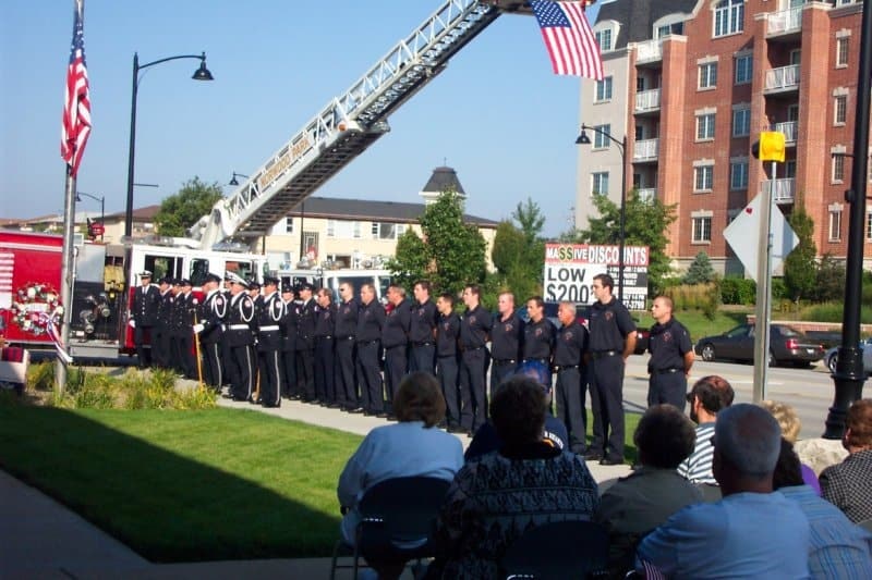 A group of firefighters stands in formation by a firetruck with a raised ladder. An audience is seated, and flags are displayed.