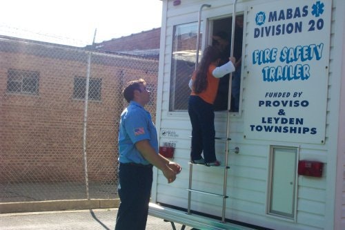 A child climbs into a fire safety trailer while an adult assists, part of a fire safety educational setup.