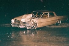 A damaged car with a crumpled front, likely involved in an accident, on a wet road at night.