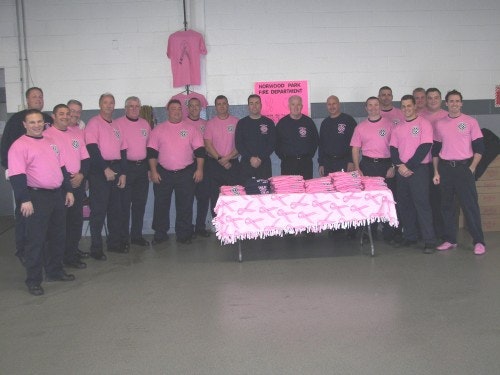 A group of people in pink shirts stand around a table with pink items, under a sign reading "Hickory Park Fire Department."