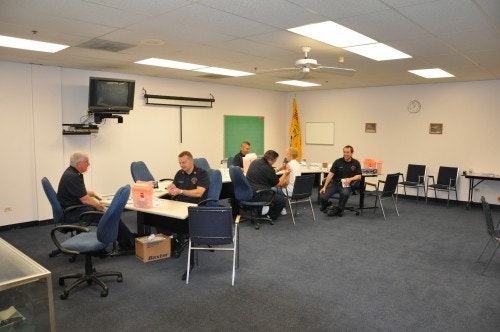 A group of people in a meeting room with tables, chairs, a TV, a wall flag, and boxes on the floor.