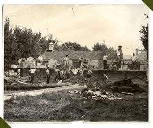 A group of people are constructing a building with bricks outdoors, surrounded by trees and materials on the ground.