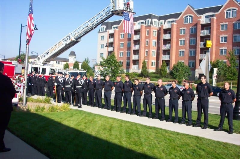A group of firefighters stands in a line for a ceremony, with a fire truck and American flags in the background.
