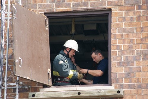 Two people are in a building window, one wearing a helmet, possibly involved in a safety or rescue operation.