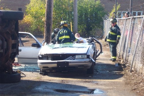 Firefighters attend to a person in a car crash scene with a flipped vehicle nearby.