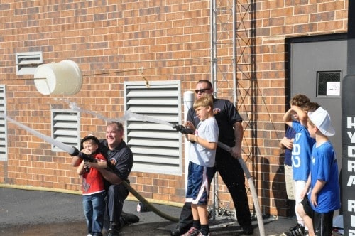 Children and adults spraying water using fire hoses at targets against a brick wall.