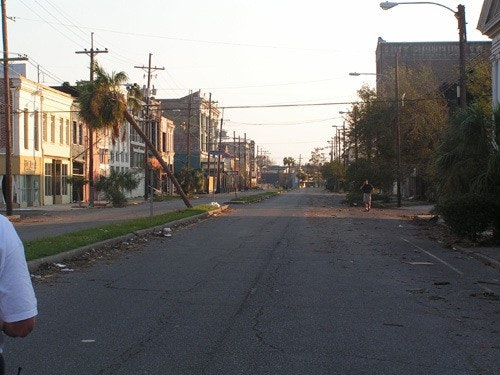 An empty street with debris and downed power lines, lined with buildings and palm trees under a clear sky.