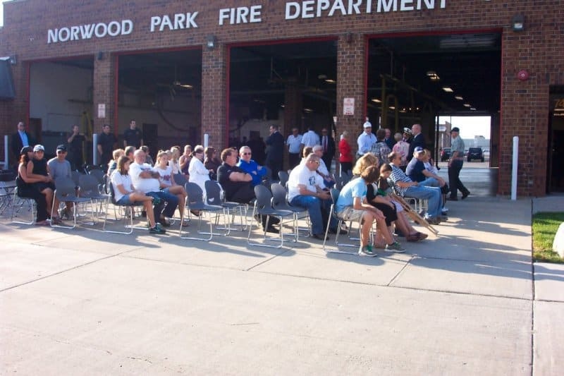 A group of people sitting and standing outside the Norwood Park Fire Department, with chairs arranged on the pavement.