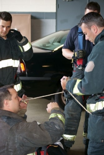 Firefighters in gear practicing a rope technique indoors.