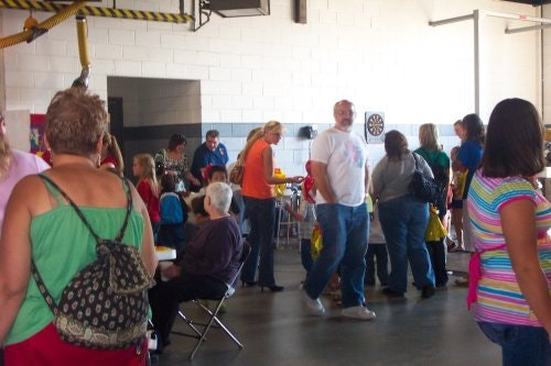 A group of people socializing indoors, some standing and others seated. There's a dartboard on the wall in the background.