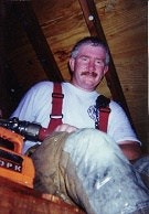 A person wearing a t-shirt and suspenders, holding a tool, inside a wooden structure.