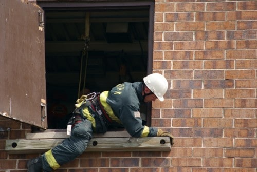 A firefighter in gear is climbing out of a window on a brick building.