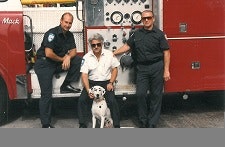 Three men pose with a Dalmatian in front of a fire truck.