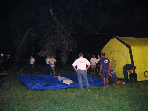 People setting up a yellow tent and inflating a blue structure at night on a grassy area, surrounded by trees.