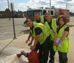 Four people in safety vests by a hydrant with water flowing. Fire truck in the background.