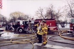 Firefighters standing near fire trucks with hoses on the ground, in a winter setting.