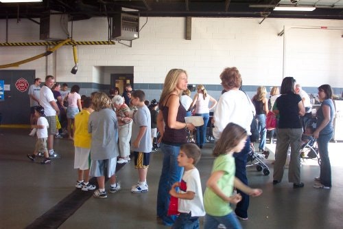 A group of people, including children and adults, are gathered indoors, possibly at an event or waiting area.