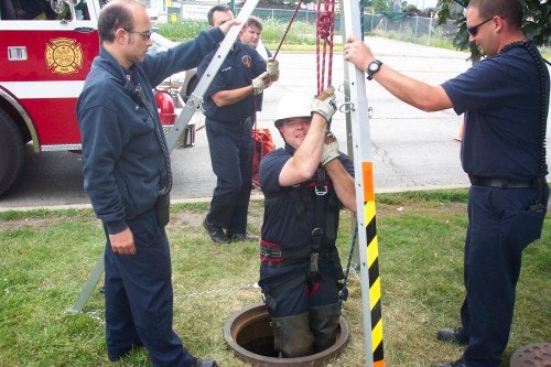 Rescue personnel are using a tripod and ropes to assist someone emerging from a manhole near a fire truck.